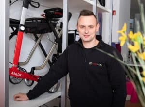 Eine Person in einer schwarzen Jacke steht neben einem roten Kinderwagen mit der Aufschrift „ATHLON“ auf einem Ausstellungsregal. Im Hintergrund sind weitere Kinderwagen und eine verschwommene Pflanze mit gelben Blüten im Vordergrund zu sehen.