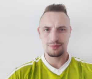 Eine Person mit kurzen Haaren und Spitzbart trägt ein limettengrünes Sporttrikot mit weißem Besatz. Der Hintergrund ist schlicht weiß. Die Person blickt mit neutralem Gesichtsausdruck direkt in die Kamera.