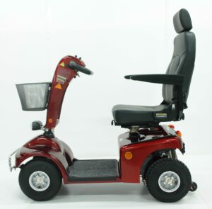 Roter Elektromobil mit schwarz gepolstertem Sitz und Armlehnen, seitlich dargestellt. Er verfügt über einen Korb vorne und vier Räder. Der Lenker ist mit Bedienelementen ausgestattet. Der Roller steht vor einem schlichten weißen Hintergrund.
